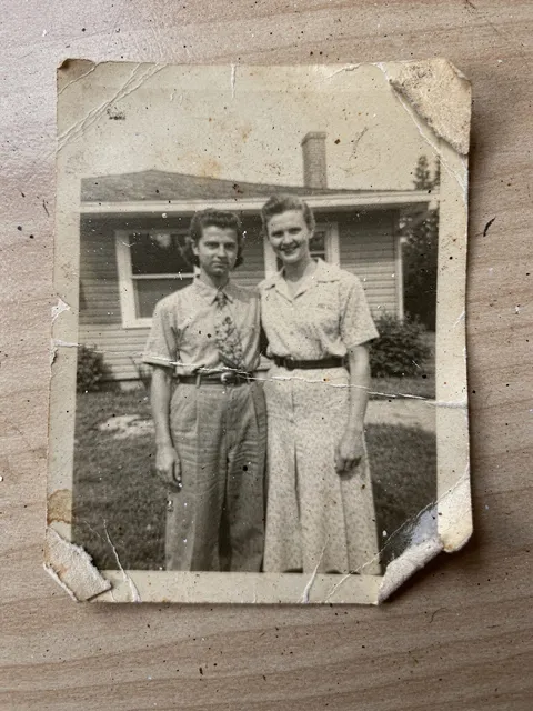 A creased, scratched black-and-white snapshot of two people in front of a house.